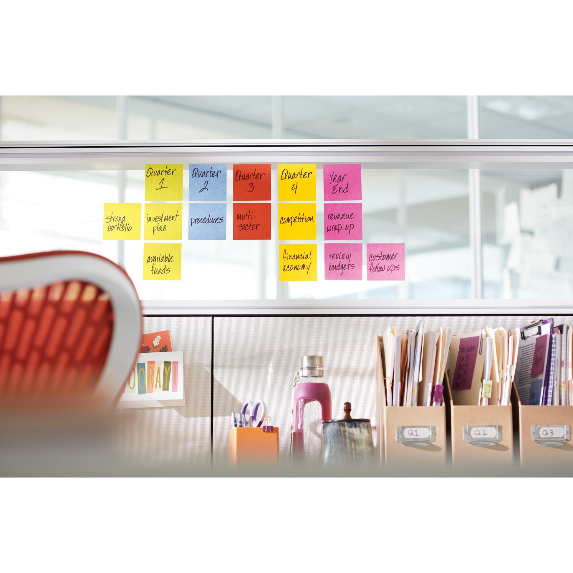 Ein Satz Post-it® Super Sticky Notes (47,6 mm x 47,6 mm, 90 Blatt/Block, 100% PEFC) der 3M Deutschland GmbH ist an einer Glaswand über einem Bürotisch mit Ordnern, einer Wasserflasche und Verbrauchsmaterialien angebracht.