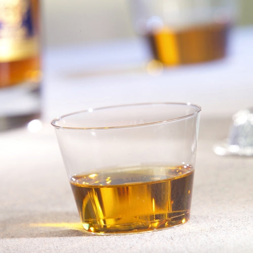 Ένα ποτήρι Starpak 15, PS 0,1 λ διαυγές, περιέχει μια μικρή ποσότητα κεχριμπαρένιου υγρού σε μια φωτεινή επιφάνεια, ενώ στο φόντο ένα άλλο ποτήρι και ένα μπουκάλι είναι θολά.