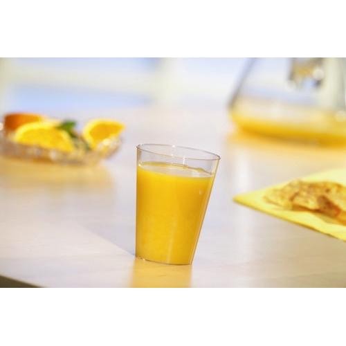 Ένα ποτήρι Starpak 40, PS 0,1 l Ø 5,5 cm · 7,5 cm διαφανές μιας χρήσης είναι γεμάτο με χυμό πορτοκαλιού πάνω σε ένα φωτεινό τραπέζι. Στο φόντο υπάρχουν φέτες πορτοκαλιού και μια κανάτα - ιδανικό για πάρτι και εκδηλώσεις.