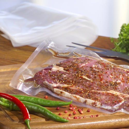 Ένας Starpak 100 Siegelrandbeutel, PA / PE διαφανής, γεμάτος με μαριναρισμένο κρέας, βρίσκεται πάνω σε ένα ξύλινο σανίδι κοπής με ολόκληρες πιπεριές τσίλι και κόκκους πιπεριού, που είναι διάσπαρτοι κοντά, και αποτυπώνει μια φρέσκια σκηνή κενού τροφίμων από Starpak.