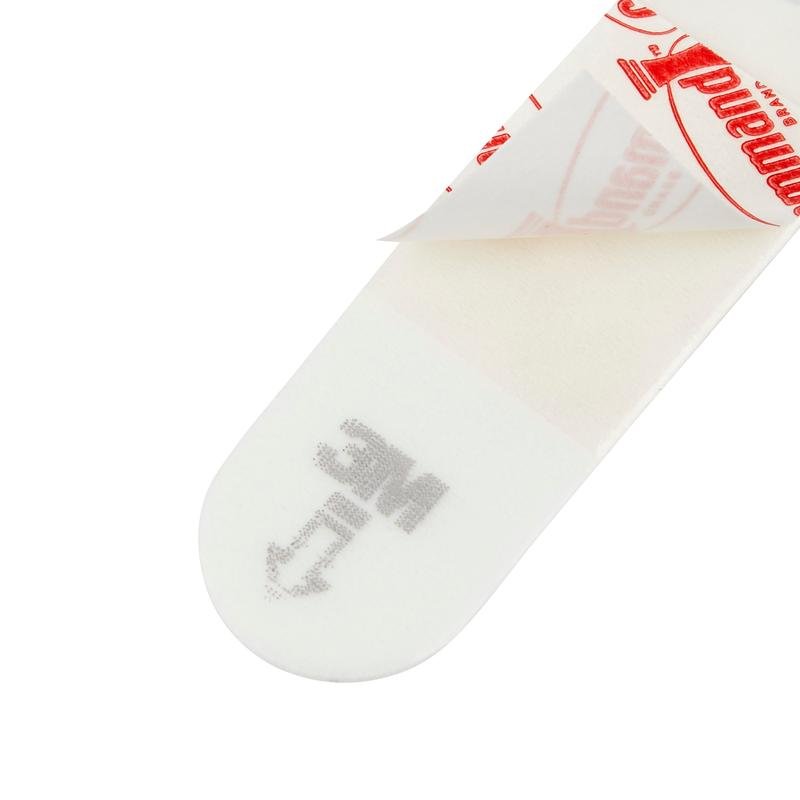 Μια κοντινή λήψη ενός Kleine Command™ Strips S, 17022N από την 3M Deutschland GmbH δείχνει το ορθογώνιο σχήμα του, με το ένα άκρο του προστατευτικού χαρτιού να έχει αφαιρεθεί και η κόλλα να αποκαλύπτεται. "MC" και ένα βέλος είναι τυπωμένα για να διευκολύνουν τη χρήση στον τοίχο χωρίς ζημιές.