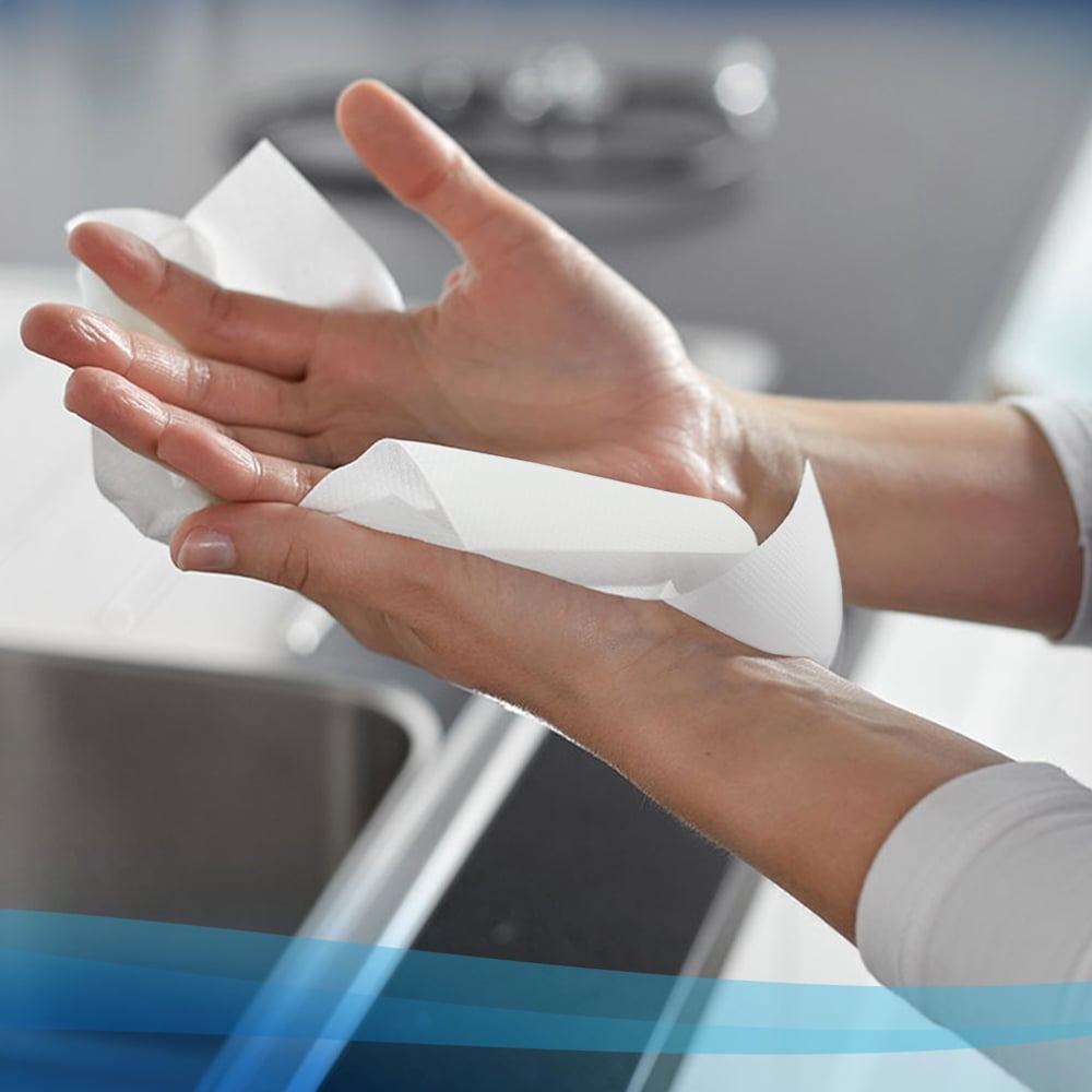 Eine Person trocknet sich die Hände mit Scott® Control™ Slimroll™ Handtüchern von Kimberly-Clark GmbH über einer Küchenspüle, mit einer unscharfen Arbeitsplatte und einem Wasserhahn im Hintergrund für hygienisches Händetrocknen.