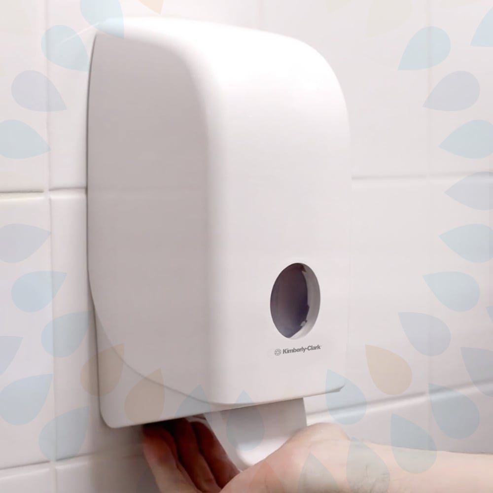 Eine Kleenex® Sanfte Waschlotion-Kassette der Kimberly-Clark GmbH (transparent, 1 L) ist auf einer weiß gekachelten Wand abgebildet, mit einer Hand, die ein Handtuch aus dem Boden zieht und einem kleinen runden Fenster auf der Vorderseite. Erhältlich in Packungen mit 6 Kassetten.