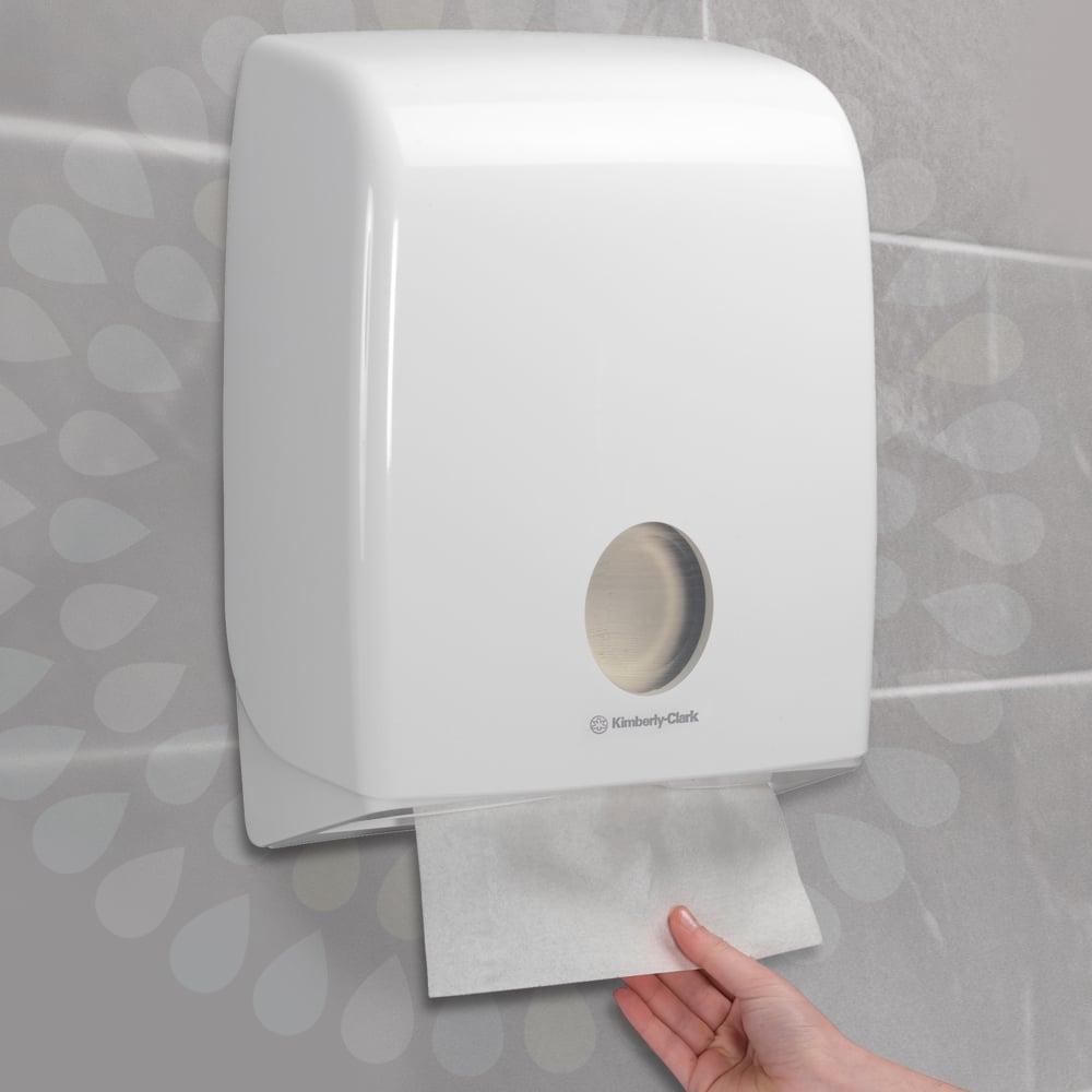 Eine Hand zieht ein Kleenex® Ultra™ Handtuch aus einem an der Wand montierten weißen Spender der Kimberly-Clark GmbH, der an einer gefliesten Wand installiert ist (Interfold/Weiß/Groß - 30 Clips x 94 Blatt | Karton, 30 Päckchen).