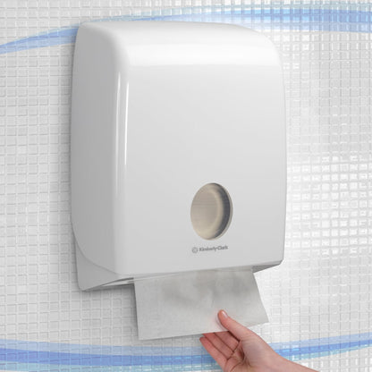 Eine Hand zieht ein Papierhandtuch aus einem an der Wand montierten, weißen Aquarius™ Spender für gefaltete Handtücher mit C-Falzung von Kimberly-Clark GmbH, installiert an einer gefliesten Wand.