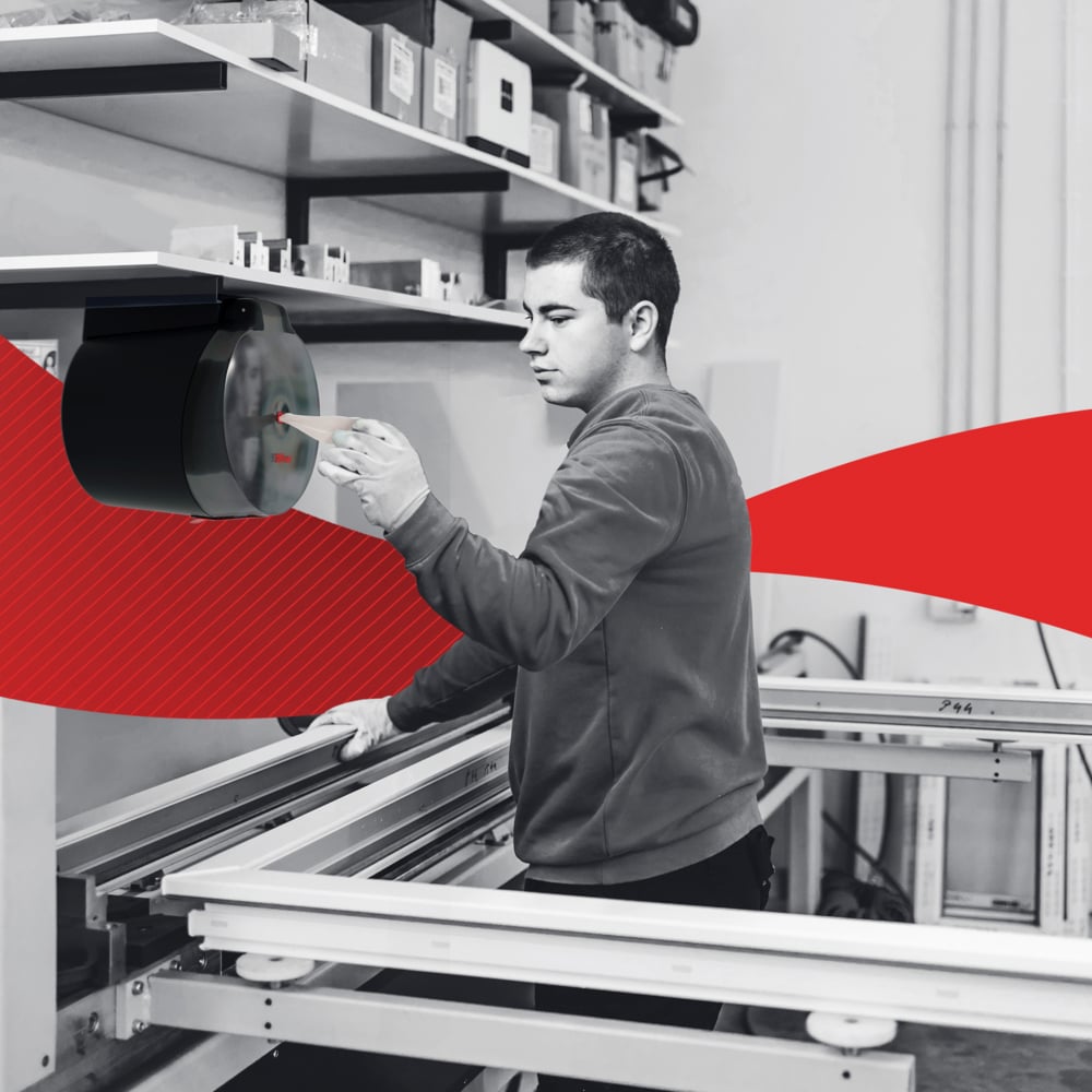 Σε ένα εργαστήριο, ένα άτομο αφαιρεί μια WypAll® L20 χαρτοπετσέτα από την Kimberly-Clark GmbH μέσω κεντρικής αφαίρεσης από τον τοίχο και καθαρίζει βιομηχανικές επιφάνειες εργασίας. ένα κόκκινο γραφικό στοιχείο διατρέχει το κέντρο της εικόνας.