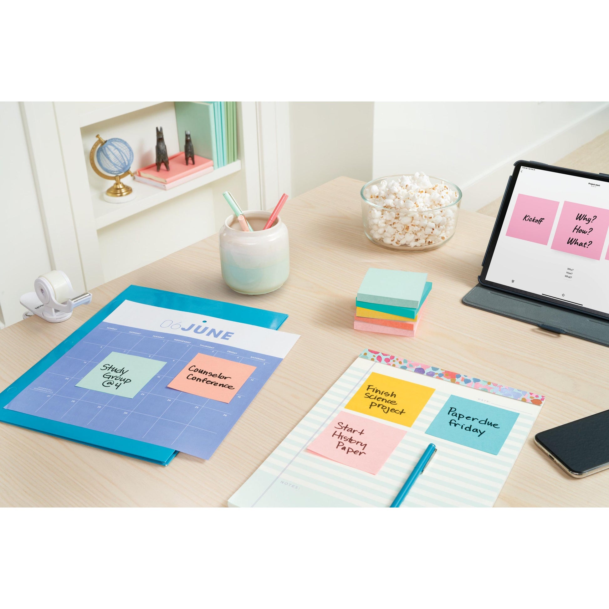 Ein organisierter Schreibtisch zum Lernen oder Arbeiten mit Post-it® Notes (76 mm x 127 mm, 100 Blatt/Block, 6 Blöcke/Packung, 100% PEFC) der 3M Deutschland GmbH, Notizbüchern, Stiften in einer Tasse, Popcorn, Klebeband, einem Tablet mit Notizen; ein Bücherregal mit Büchern schmückt den Hintergrund.