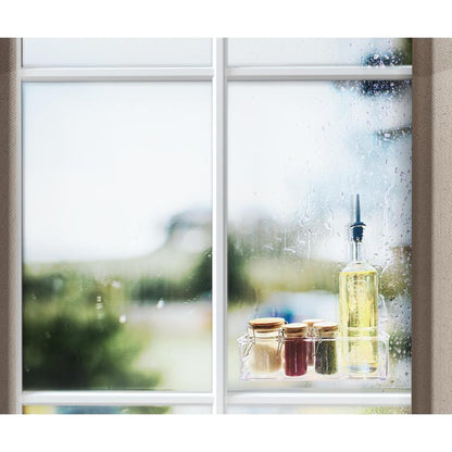 Ένα παράθυρο με σταγόνες βροχής δείχνει μια σκηνή εξωτερικού χώρου. Στο περβάζι παραθύρου βρίσκεται μια φιάλη λαδιού, τρία μικρά γυάλινα δοχεία με χρυσά καπάκια και ένα ρολό διάφανης διπλής όψης ταινίας Scotch-FIX™ 4910C της 3M Deutschland GmbH.