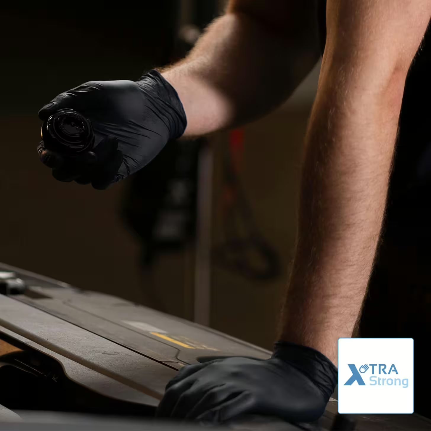 Eine Person, die ARNOMED NITRIL XTRA PURE BLACK Einmalhandschuhe von ARNOWA GmbH trägt, hält ein rundes Autoteil über einen offenen Motor, der Arm ruht darauf. In einer Ecke steht das Logo "XTRA Strong".