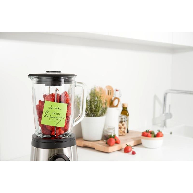 Ένα μπλέντερ με φράουλες βρίσκεται σε μια λευκή κουζίνα. Ένα Post-it® Super Sticky Note (76 x 76 mm, 90 φύλλα, 100 % PEFC) της 3M Deutschland GmbH είναι προσαρτημένο στο δοχείο. Κοντά βρίσκονται σκεύη, φυτά σε γλάστρες, ελαιόλαδο και άλλες φράουλες σε ένα ξύλο κοπής.