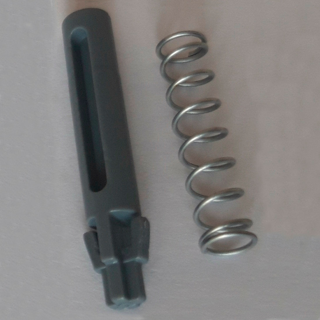 Ένα μεταλλικό ελατήριο και ένας σκούρος γκρι κυλινδρικός εφεδρικός πείρος πόρτας με αυλακώσεις και δόντια, μέρος του DI Protect Türzapfen & Feder (1 Stück) από την Diversey Deutschland GmbH & Co. OHG, απεικονίζονται σε μια φωτεινή επιφάνεια και δείχνουν τα ανθεκτικά υλικά για το DI Protect Trolley.