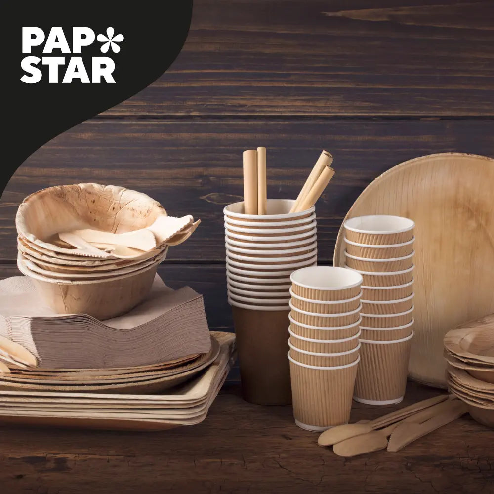 Verschiedene biologisch abbaubare Einweggeschirre, darunter Pappteller, -becher, -schüsseln, -servietten und Holzbesteck, angeordnet auf einer hölzernen Fläche mit einem 