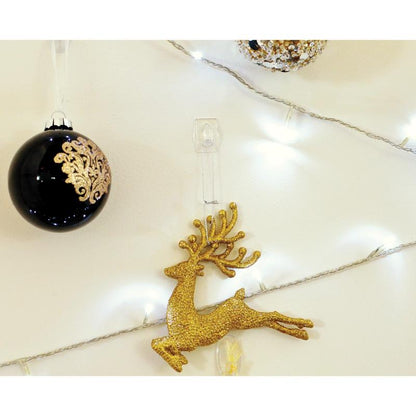 Ένα χρυσό γκλίτερ στολίδι τάρανδου κρέμεται από ένα Command™ Mini-Haken, transparent, mit transparenten Strips (17006CLRFGN) von 3M Deutschland GmbH, με μια μαύρο-χρυσή χριστουγεννιάτικη μπάλα και λευκό χριστουγεννιάτικο φωτισμό στο φόντο.