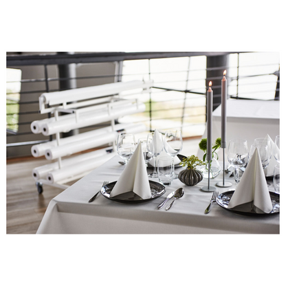 Σε ένα όμορφα στρωμένο τραπέζι δείπνου υπάρχει ένα φρέσκο, λευκό ρολό τραπεζομάντηλου Duni Dunicel® της Duni GmbH, που προσφέρει υφή που μοιάζει με ύφασμα. Τριγωνικά διπλωμένες χαρτοπετσέτες σε σκούρα πιάτα συνοδεύουν ποτήρια κρασιού. Ψηλά κεριά και μικρές ανθοσυνθέσεις χρησιμεύουν ως διακόσμηση τραπεζιού, στο φόντο φαίνονται τυλιγμένοι καμβάδες.