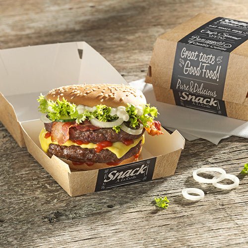 Ένα cheeseburger με μαρούλι, ντομάτα, μπέικον, κρεμμύδια και σάλτσα βρίσκεται σε ένα ανοιχτό PAPSTAR 50 κουτί burger από χαρτόνι "pure" της PAPSTAR GmbH πάνω σε ξύλινο τραπέζι. Δίπλα ένα κλειστό κουτί με εκτύπωση "Good Food" και Onion Rings.