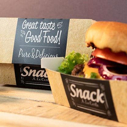 Ένας δίσκος από χαρτόνι με την επιγραφή "Snack A La Carte" περιέχει ένα χάμπουργκερ με μαρούλι, ντομάτα και κρεμμύδι. Στο παρασκήνιο υπάρχει μια PAPSTAR 50 κουτιά burger, χαρτόνι "pure" "Good Food" της PAPSTAR GmbH: Καλή γεύση Good Food! Καθαρό & Νόστιμο.