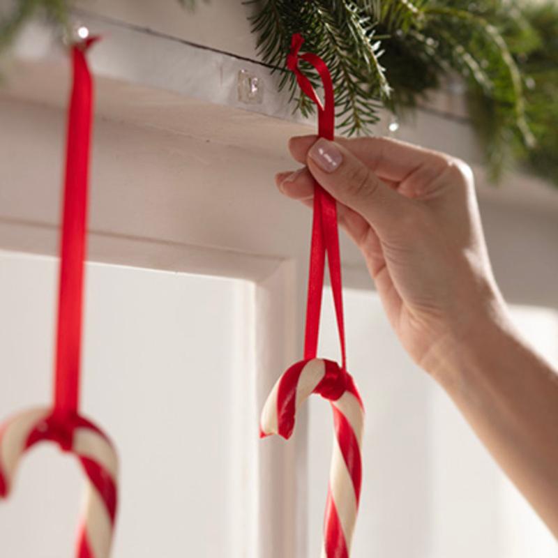 Ένα χέρι κρεμάει μια κόκκινη-λευκή καραμέλα σε κόκκινη κορδέλα σε λευκή επιφάνεια, με Command™ Mini-Haken, transparent, mit transparenten Strips, 17006CLRFGN von 3M Deutschland GmbH. Ένα άλλο ζαχαρωτό κρέμεται ήδη δίπλα σε πράσινη γιρλάντα έλατου.