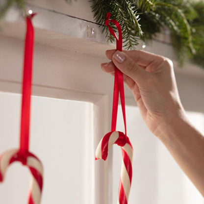 Ένα χέρι κρεμάει μια κόκκινη-λευκή καραμέλα σε κόκκινη κορδέλα σε λευκή επιφάνεια, με Command™ Mini-Haken, transparent, mit transparenten Strips, 17006CLRFGN von 3M Deutschland GmbH. Ένα άλλο ζαχαρωτό κρέμεται ήδη δίπλα σε πράσινη γιρλάντα έλατου.