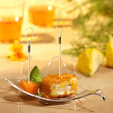 Ένα γυάλινο μπολ παρουσιάζει έναν πανέ, τηγανισμένο κύβο ορεκτικού με έναν PAPSTAR Fingerfood Picker "Loop" (10 cm), ένα τμήμα πορτοκαλιού και μία πράσινη γαρνιτούρα. Στο υπόβαθρο, κομμάτια λεμονιού και δύο ποτά της PAPSTAR GmbH θολώνουν.