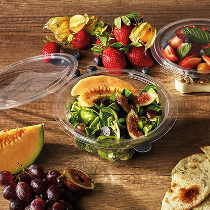 Μια σαλατιέρα Starpak 50 με καπάκι, R-PET διαφανής στρογγυλή, περιέχει σαλάτα πεπονιού και σύκων σε ξύλινο τραπέζι με φράουλες, σταφύλια, ψωμί, κεράσια και φρούτα. Ένα άλλο δοχείο Starpak R-PET με φρούτα βρίσκεται κοντά.