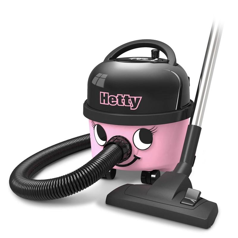 Η ηλεκτρική σκούπα Hetty Compact HET160-11 της Numatic International GmbH είναι μια ροζ-μαύρη σκούπα καναλιού με ένα χαμογελαστό πρόσωπο, έναν ευέλικτο σωλήνα, μια κινητή βάση και μια κεφαλή απορρόφησης που στέκεται δίπλα στη συσκευή στο πάτωμα.