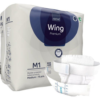 Μια συσκευασία Abena Re-Seller GmbH ABENA Wing Premium απεικονίζεται με ένα ανοιγμένο μαξιλάρι μπροστά που προσφέρει προστασία και άνεση. Στη συσκευασία αναφέρεται ότι περιέχει 15 μεσαίου μεγέθους μαξιλάρια για προστασία της ουροδόχου κύστης.