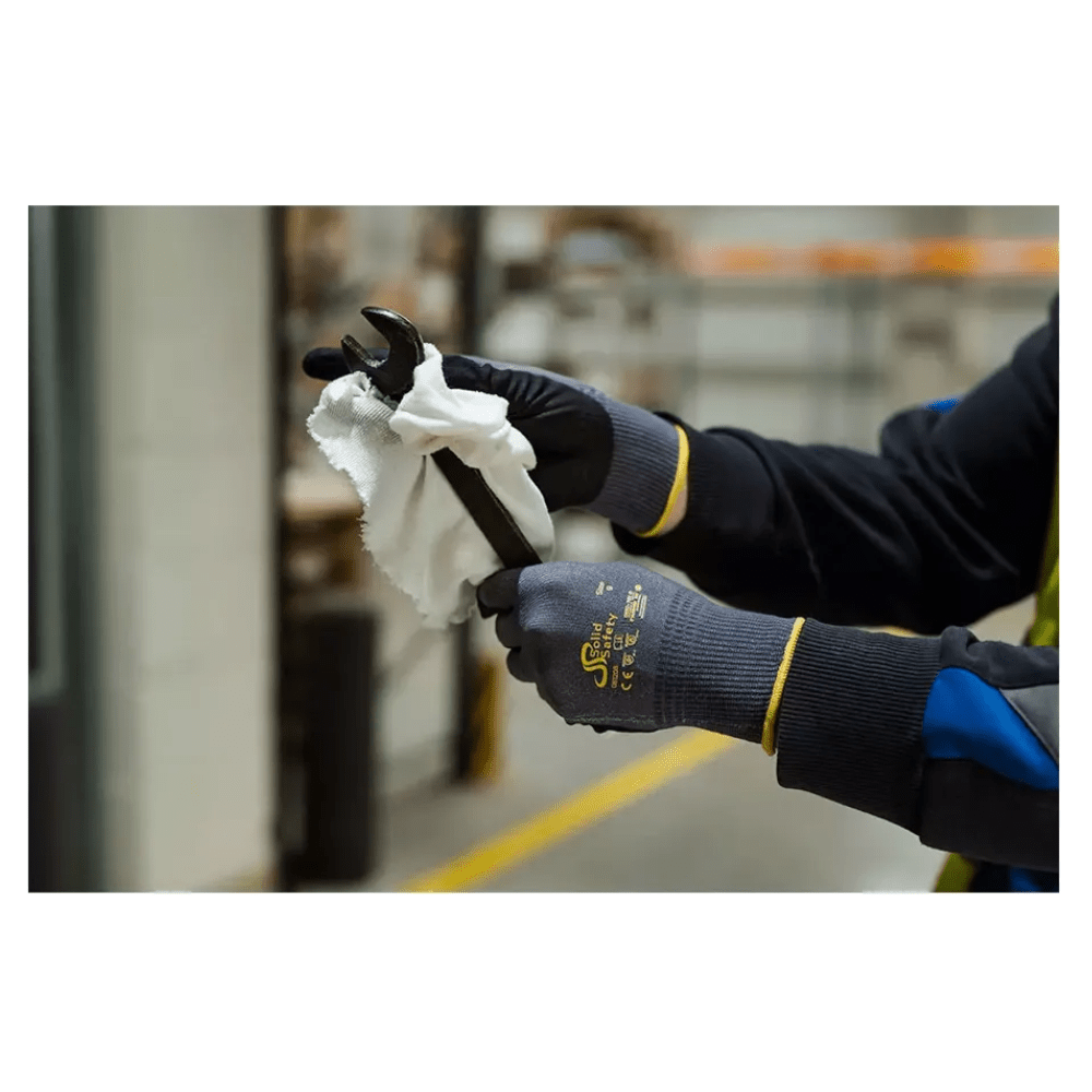 Ένα άτομο με AMPri SolidSafety Tough Air υπερελαφριά γάντια συναρμολόγησης της AMPri Handelsgesellschaft mbH καθαρίζει προσεκτικά έναν κλειδί με ένα λευκό πανί σε εσωτερικό χώρο. Τα γκρι γάντια, που είναι γνωστά για το επίπεδο αντοχής στην τριβή 4, ξεχωρίζουν από τα θολά βιομηχανικά ράφια και συσκευές στο παρασκήνιο.