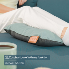 Ένα άτομο κάθεται σε έναν καναπέ, τα πόδια του είναι τεντωμένα και ξεκουράζονται σε ένα μπλε-γκρι Beurer μαξιλάρι μασάζ με μπαταρία MG 139 της Beurer GmbH. Το μαξιλάρι έχει μια μικρή ανοιχτόχρωμη καφέ ετικέτα και ένα αυτοκόλλητο με την επιγραφή «Λειτουργία θερμότητας με δύο επίπεδα», που υποδεικνύει ότι το μαξιλάρι διαθέτει ρυθμιζόμενες λειτουργίες θερμότητας.