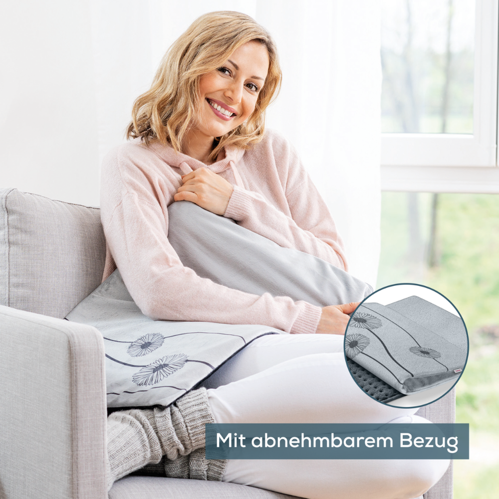 Eine Frau sitzt auf einer hellgrauen Couch neben einem Fenster und hält das Beurer HK 125 XXL-Heizkissen Cosy Grey der Beurer GmbH in der Hand. Sie lächelt und trägt einen rosa Kapuzenpullover. Es gibt eine Nahaufnahme des Kissens, die seinen abnehmbaren Bezug und die Heizfunktionen mit 6 Wärmestufen zeigt. Der deutsche Text lautet „Mit abnehmbarem Bezug.