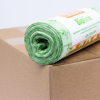 Ένας ρόλος πράσινων σακουλών βιοαποβλήτων τοποθετείται σε ένα χαρτόνι. Η ετικέτα στον ρόλο είναι εν μέρει ορατή και φέρει την εκτύπωση «EMIL DEISS KG (GmbH + Co.)». Αυτές οι 100% κομποστοποιήσιμες σακούλες έχουν ένα λουλουδάτο σχέδιο και είναι 430 x 500 mm μεγάλες.