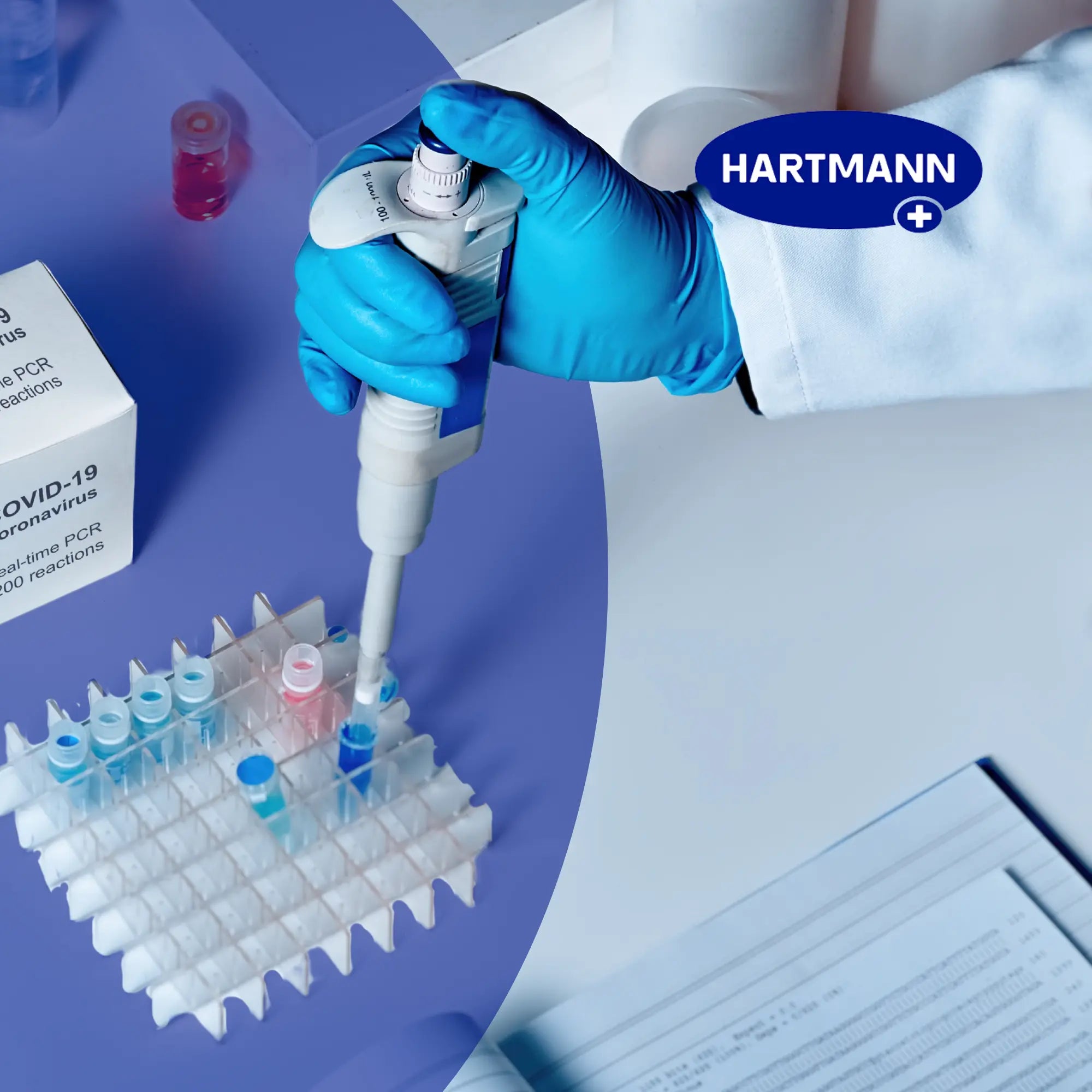 Eine behandschuhte Hand benutzt eine Pipette, um in einem Labor Flüssigkeit in Reagenzgläser zu füllen. Zu sehen sind ein COVID-19-Testkit, ein Reagenzglasgestell, ein Arbeitsblatt und das Wort "HARTMANN".