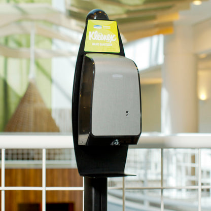 Μια ηλεκτρονική βάση λοσιόν πλυσίματος Kimberly-Clark GmbH (Μαύρο, 1 διανομέας) είναι τοποθετημένη εσωτερικά κοντά σε λευκό μεταλλικό κιγκλίδωμα για υγιεινό καθαρισμό χεριών, με θολές αρχιτεκτονικές λεπτομέρειες στο παρασκήνιο.