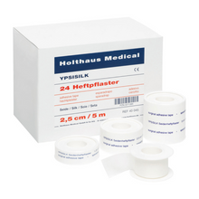 Ένα κουτί με την επιγραφή «Holthaus Medical GmbH & Co. KG» με «Holthaus YPSISILK Heftpflaster - 2,5 cm x 5 m | Συσκευασία (5 m)» και αρκετούς ρόλους λευκής κολλητικής ταινίας σε διάφορα μεγέθη μπροστά. Το κείμενο στο κουτί και στις ταινίες περιέχει λεπτομέρειες προϊόντος σε πολλές γλώσσες.