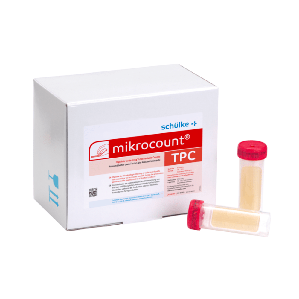 Ένα λευκό κουτί με την επιγραφή «Schülke & Mayr GmbH Mikrocount® TPC, θρεπτικό υπόστρωμα | Συσκευασία (20 τεμάχια)» με ένα λογότυπο και μια περιγραφή προϊόντος δίπλα σε δύο φιαλίδια για μικροβιακές δοκιμές, το ένα με κίτρινο καπάκι, το άλλο με διαφανές καπάκι.