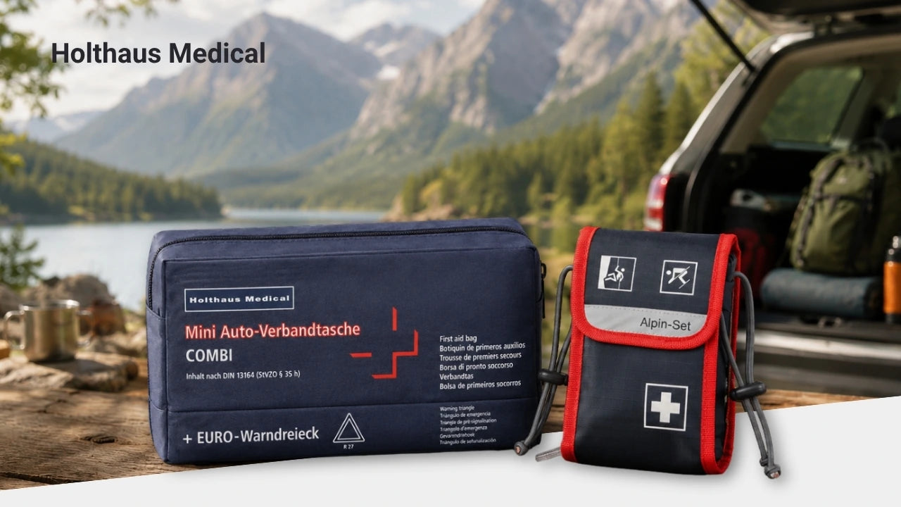 Zwei Erste-Hilfe-Sets von Holthaus Medical stehen auf einem Holztisch vor einer Berg- und Seenlandschaft. Im Hintergrund ist der geöffnete Kofferraum eines Autos mit Outdoor-Ausrüstung zu sehen.