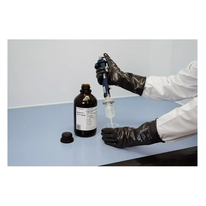Ένα άτομο που φοράει AMPri SolidSafety ChemV Full Stabile Brom-Butyl-Chemikalienschutzhandschuhe με εξωτερικό στρώμα Viton και ένα λευκό εργαστηριακό παλτό, απορροφά υγρό από μια καφέ φιάλη με μια πιπέτα σε μια ανοιχτό γκρι επιφάνεια. Το καπάκι βρίσκεται κοντά στον εργαστηριακό χώρο εργασίας και διασφαλίζει τη χημική αντοχή κατά το χειρισμό ουσιών.