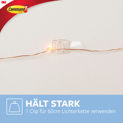 Ein Command™ Dekorations-Clip, transparent mit transparenten Strips von 3M Deutschland GmbH, hält eine Lichterkette an der Wand. Im Bild ist eine blaue Box mit deutschem Text und Command-Logo oben links zu sehen.