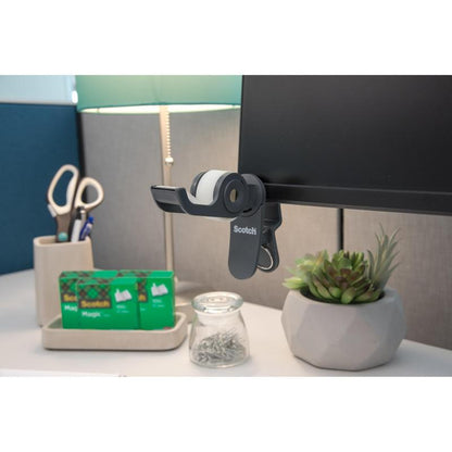 Στο γραφείο: ένας διανομέας ταινίας 3M Scotch™ Clip & Twist με αόρατη ταινία Scotch® Magic™ (19 mm × 8,89 m), μια λάμπα, ένα ψαλίδι σε μια βάση, δύο επιπλέον ρολά ταινίας Scotch®, ένα βάζο με καρφίτσες και ένα μικρό φυτό παχύφυτου σε γλάστρα.